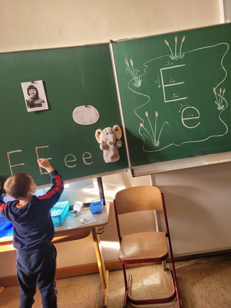 Ein Junge schreibt den Buchstaben E auf eine grüne Tafel